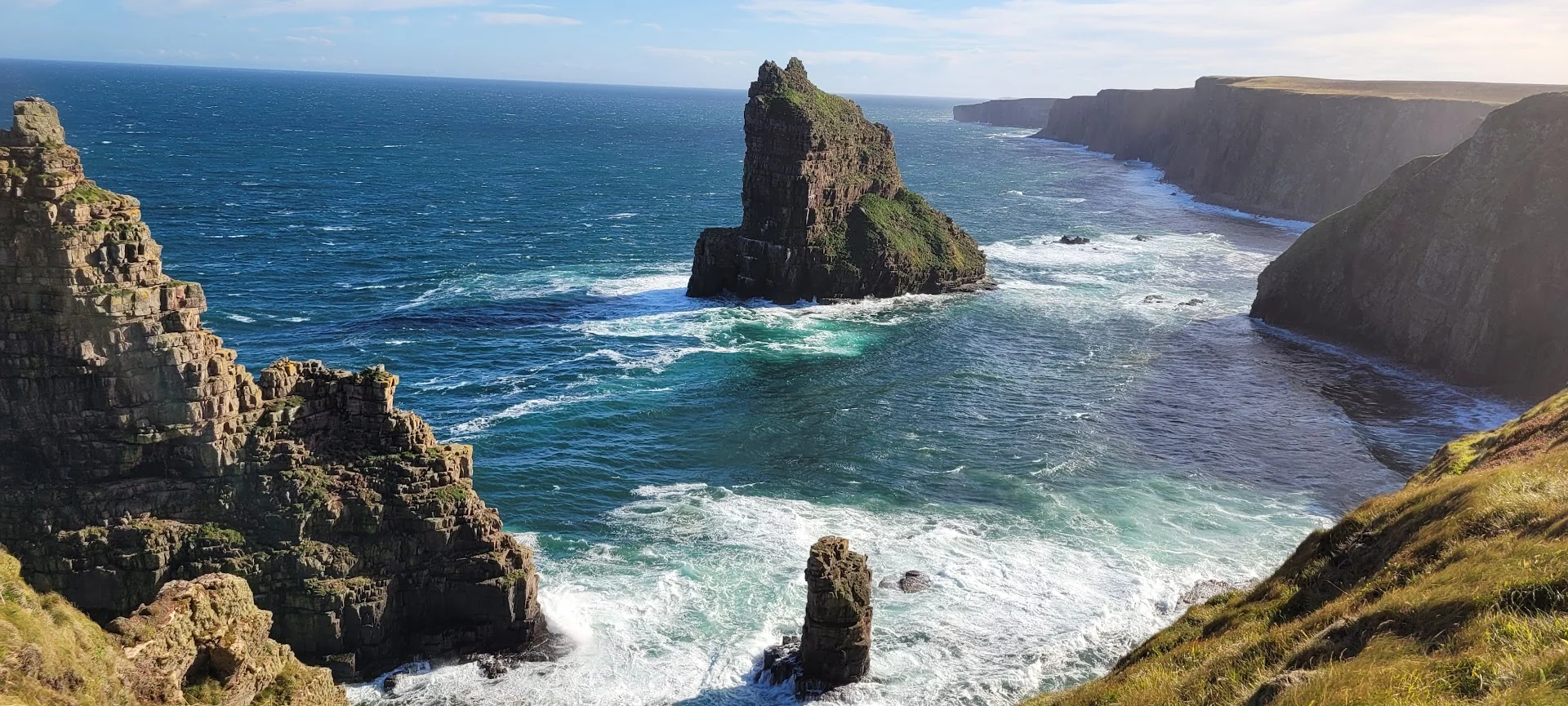 Dark sea stacks rise from turquoise Atlantic water below long, sunlit cliffs on the Irish coast.