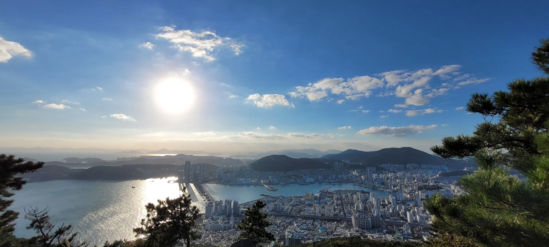 A high panoramic view over Busan, with sunlight on the bay, dense city blocks and dark mountains.