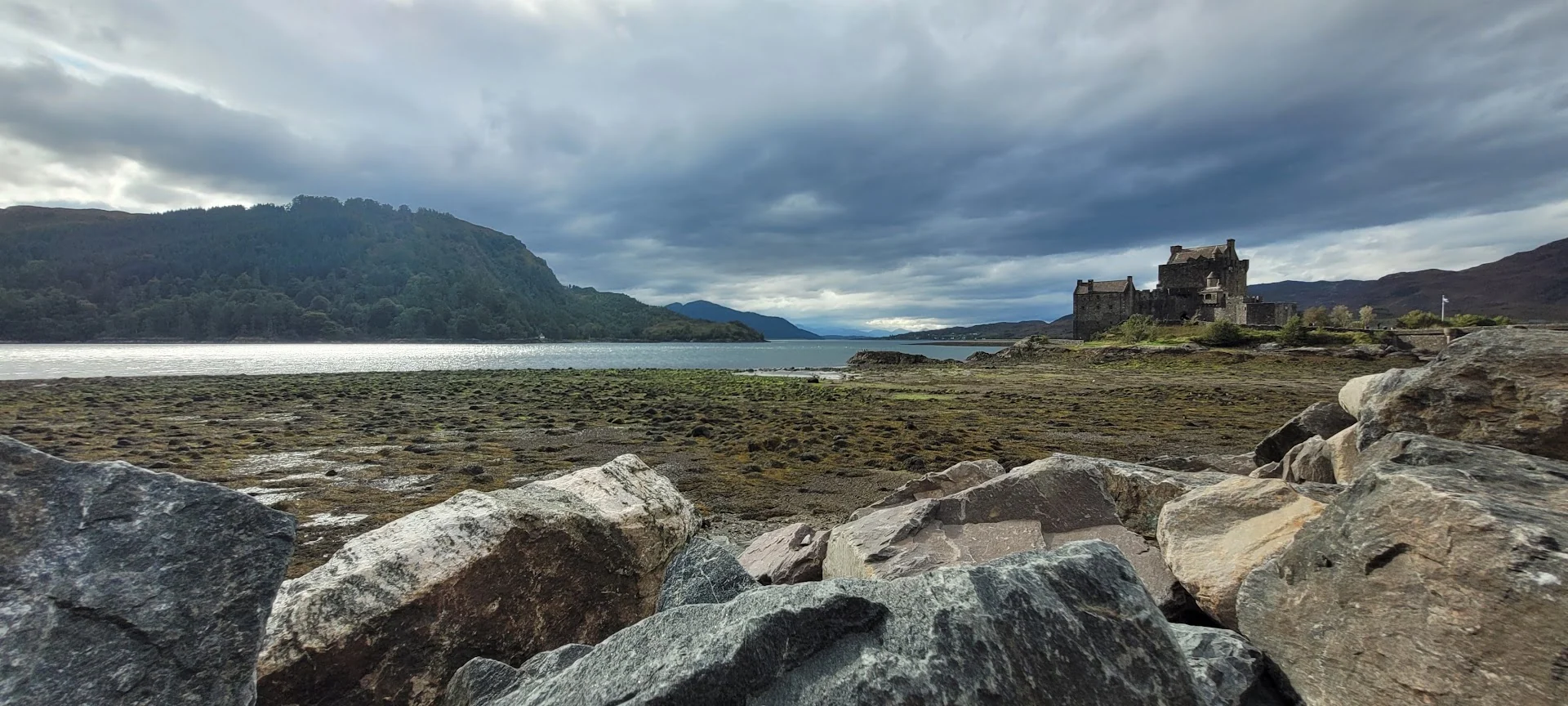 A distant stone castle stands beyond a rocky tidal shore under heavy clouds in the Scottish Highlands.
