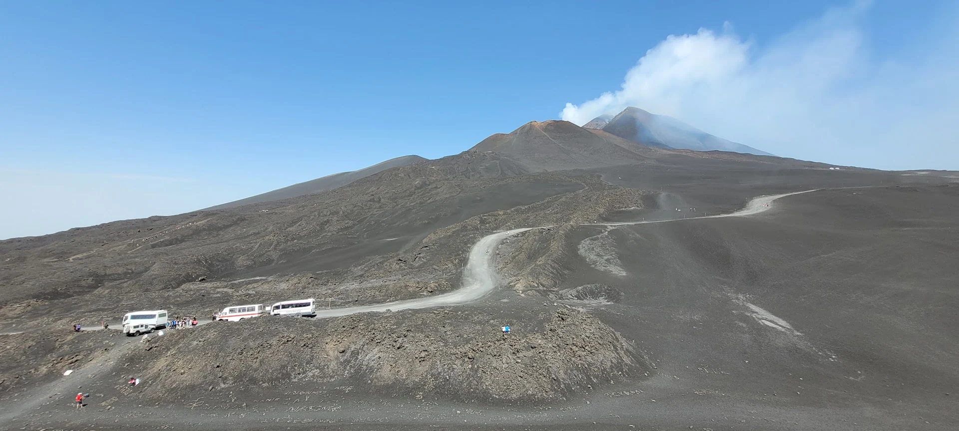 A pale road crosses black volcanic slopes toward Mount Etna, where white smoke streams from the summit.