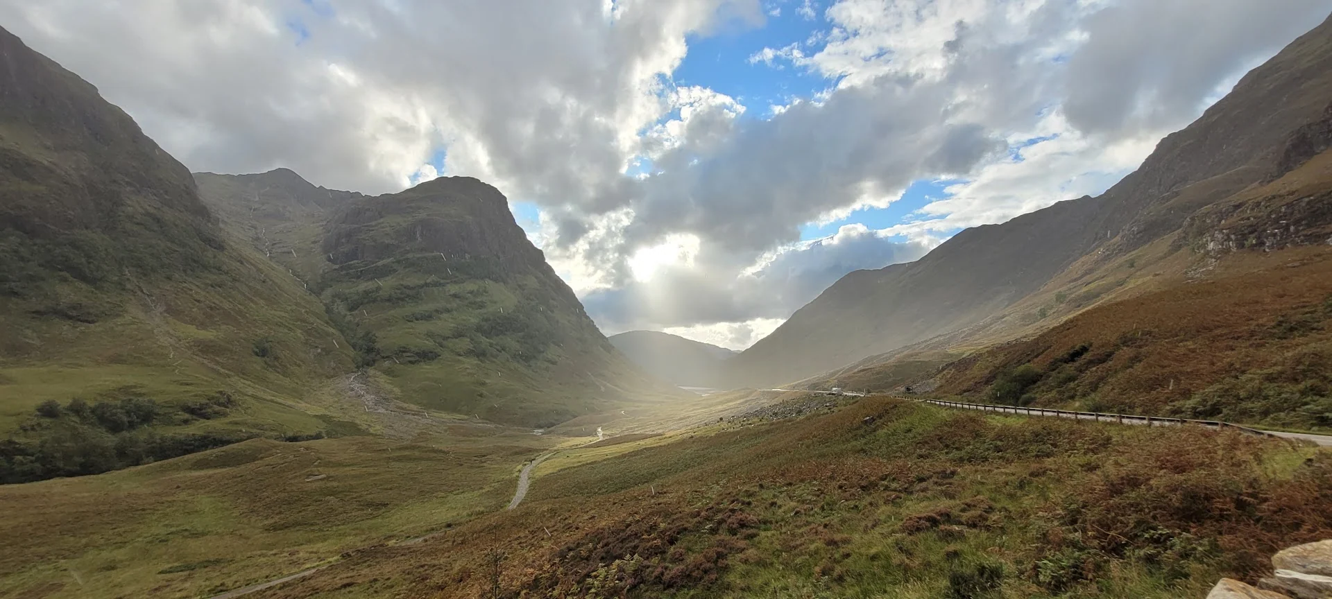 Steep green mountains form a Scottish valley as sunlight breaks through dramatic clouds onto the floor below.