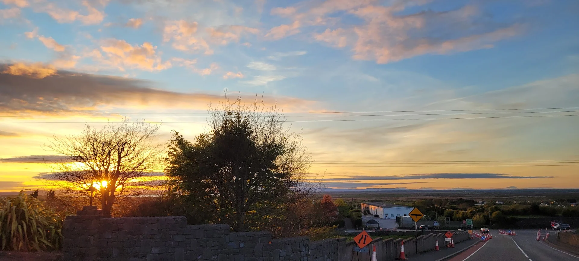 A colourful sunset spreads above trees and a Galway road descending past temporary roadworks.