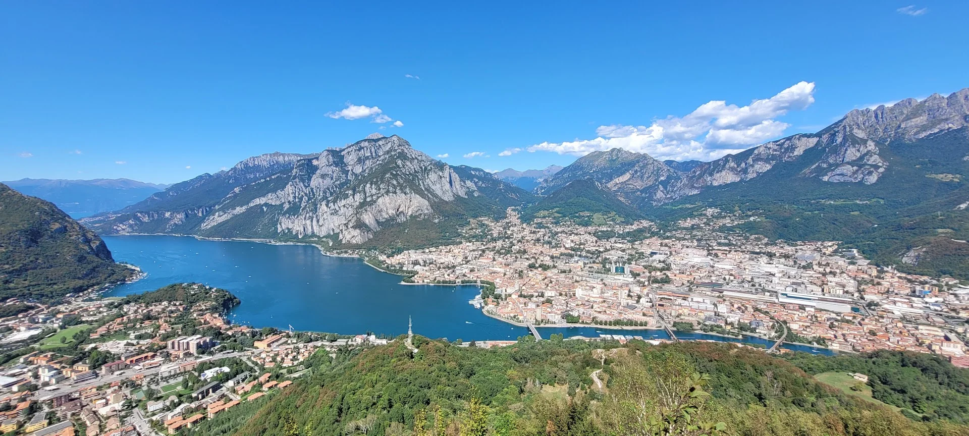 A wide elevated view of Lecco wrapped around a blue lake beneath steep green and grey mountains.
