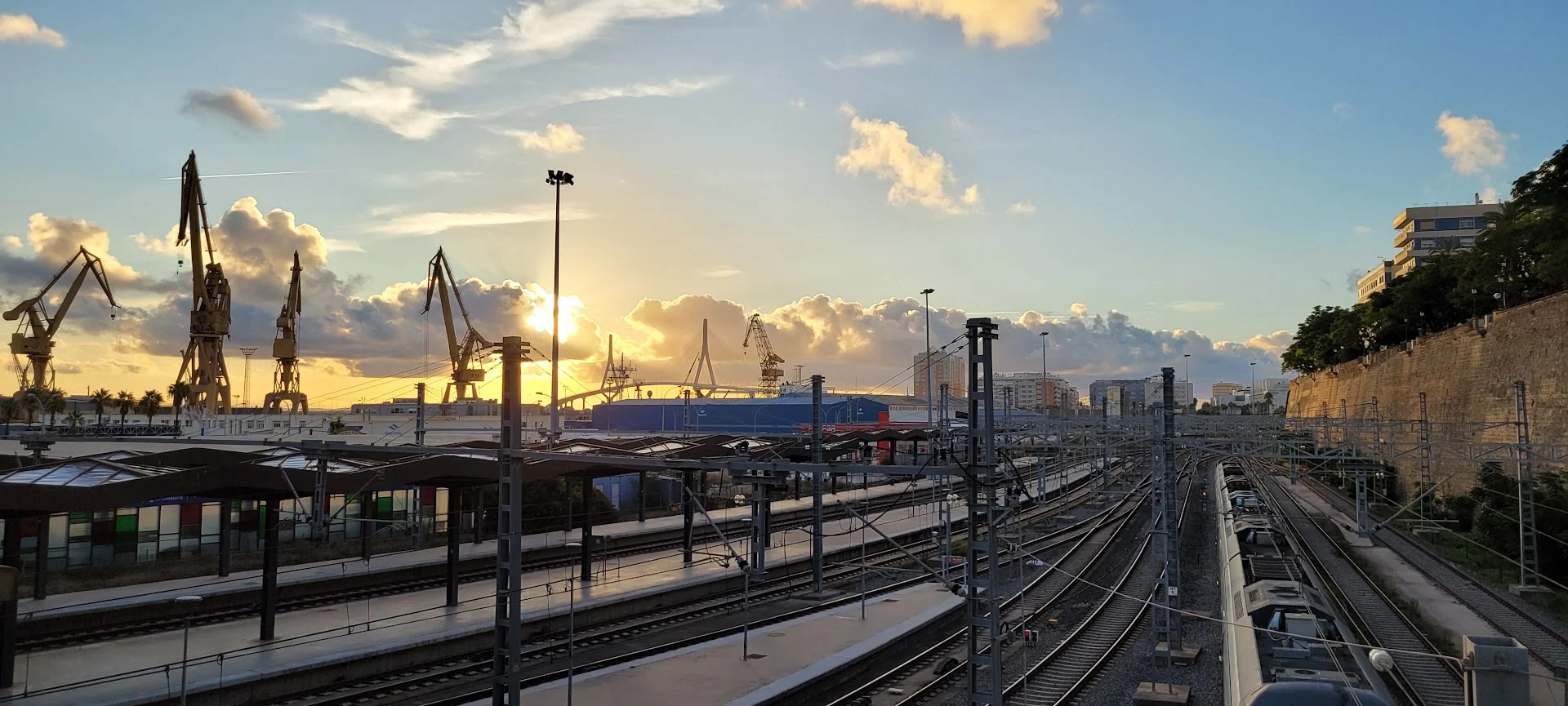 Railway tracks and overhead lines run toward a port of silhouetted cranes beneath a golden sunset.