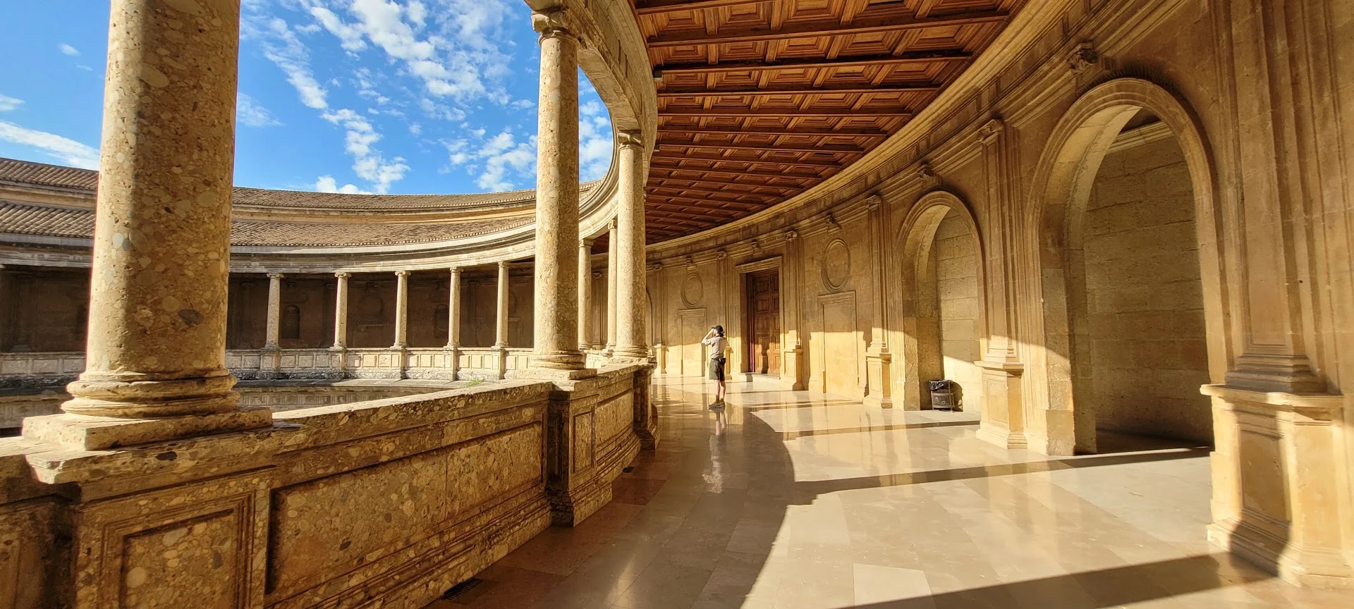 Sunlit stone columns and carved arches curve around an open courtyard beneath a blue sky.