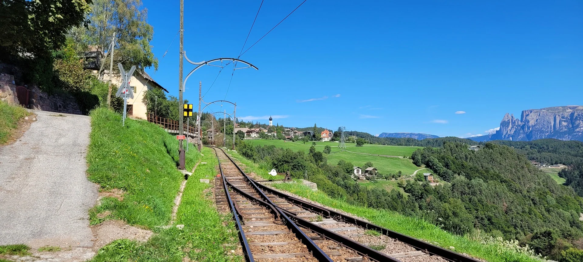 A narrow railway curves through vivid green alpine fields toward villages and sheer Dolomite cliffs.